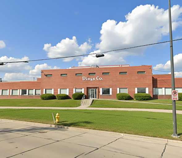 A large red brick building with the sign Dings Co. on the front, set against a blue sky with scattered clouds. There is a grassy lawn, a sidewalk, and a fire hydrant in front of the building.